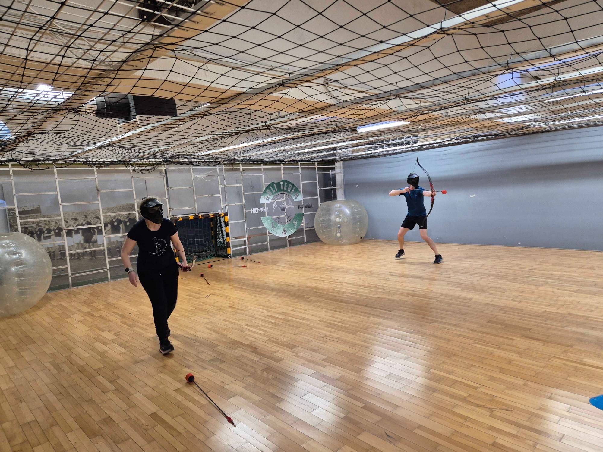 Two colleagues playing archery tag in a sports hall, one of them drawing a bow